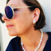 Dark haired woman wearing a blue top, large round sunglasses and Leoni & Vonk pearl jewellery. She is looking to the side of the image.