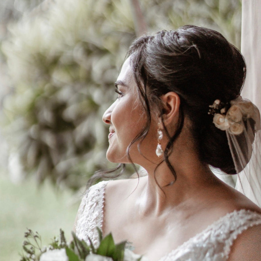 Image of a dark haired bride looking out of a window. She has her hair up and is wearing Leoni & Vonk baroque pearl and crystal bridal earrings