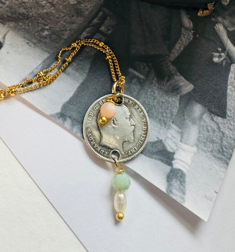 Leoni & Vonk antique 1906 threepence necklace on a gold chain on a white background and with a vintage postcard
