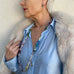 Woman leaning against a wall with her face obscured. She is wearing a blue shirt, cream fur coat and Leoni & Vonk jewellery 