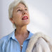 Woman with grey hair wearing a blue shirt and leoni & Vonk pearl jewellery. She is looking up to the right of the image and has a cream fur coat on her shoulder.