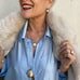 Woman wearing a blue shirt and cream fur jacket. She is holding the jacket open and wearing Leoni & Vonk vintage and antique jewellery.