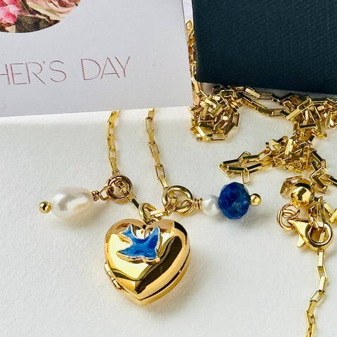 Leoni & Vonk 9ct gold plated bluebird locket with pearl and lapis charms on a gold chain. The neckalce is on a white background with the corner of a Mother's day card in the background.