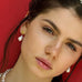 Dark haired girl wearing Leoni & Vonk gold stud and pearl earrings standing against a red wall.