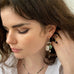Dark haired model holding her hair behind her ear wearing Leoni & Vonk jewellery and a white shirt. She is looking down to the left of the image.