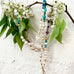 A white brick wall with a Leoni & Vonk pearl and gemstone necklace hanging from a branch with green leaves and blossom. 