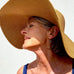 Grey haired woman wearing a large sun hat and Leoni & Vonk turqouie and pearl jewellery. She has her eys closed and is looking upwards to the left of the image.
